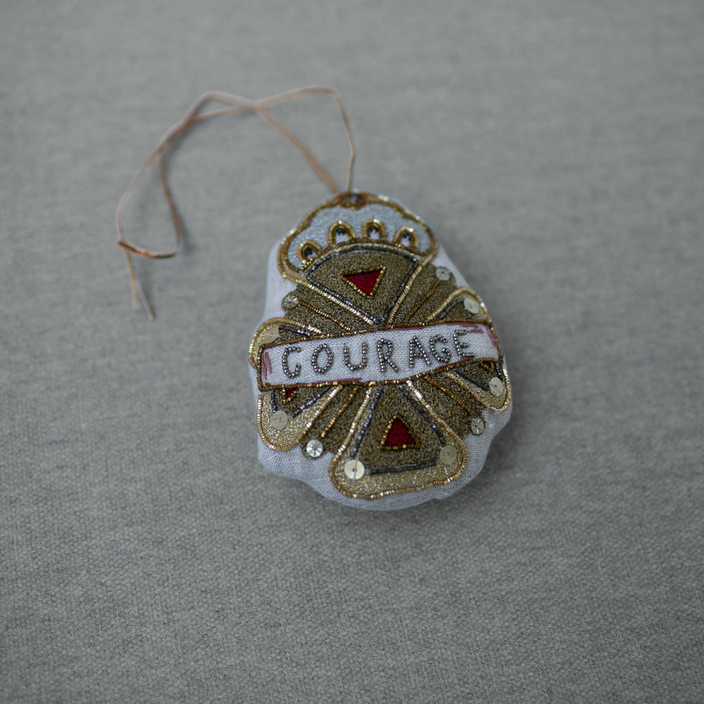 The Skippy Cotton Courage Crest Token is an Oz-inspired cotton-filled ornament, embellished in gold, red, and white with "COURAGE" at the center. It hangs by a thin string and is displayed on a gray fabric surface.