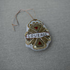 The Skippy Cotton Courage Crest Token is an Oz-inspired cotton-filled ornament, embellished in gold, red, and white with "COURAGE" at the center. It hangs by a thin string and is displayed on a gray fabric surface.
