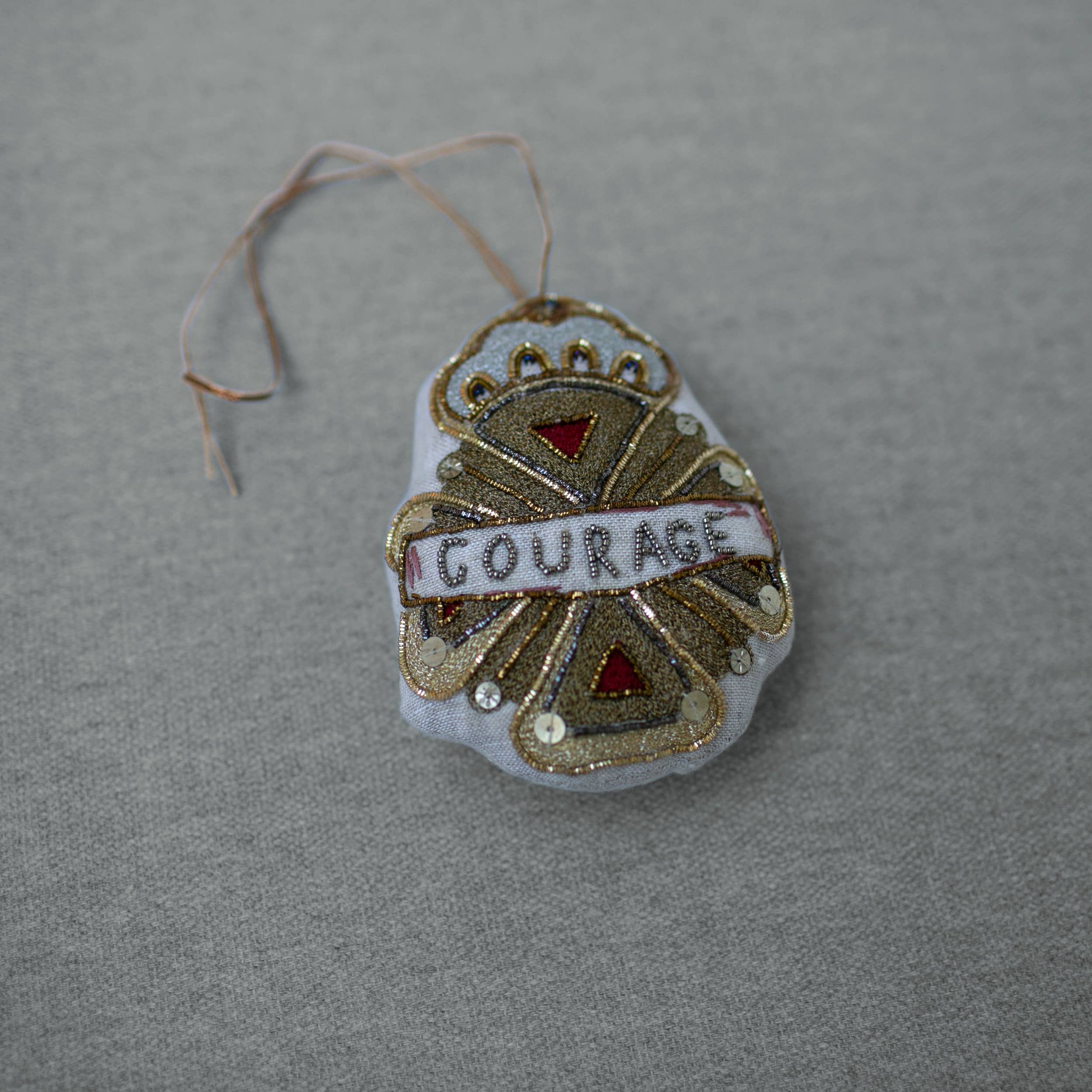 The Skippy Cotton Courage Crest Token is an Oz-inspired cotton-filled ornament, embellished in gold, red, and white with "COURAGE" at the center. It hangs by a thin string and is displayed on a gray fabric surface.