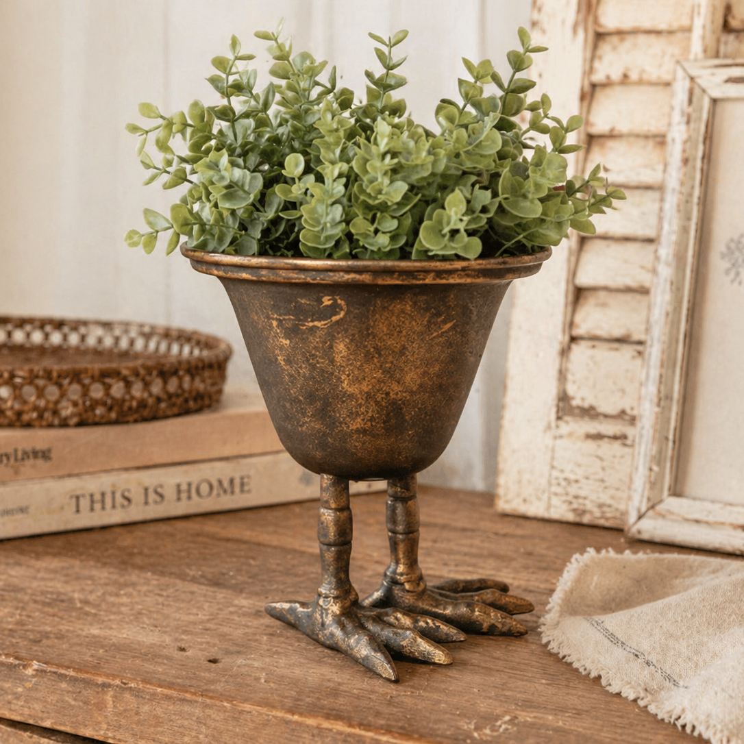 The Vagabond Vintage Metal Bird Claw Pedestal Planter, with a green leafy plant, sits on a rustic wooden table beside books and a cloth.