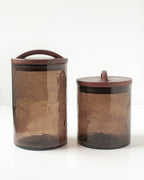 Two blush, textured Handblown Hammered Glass Canisters: Blush / Small by Creative Women with flat, hand-carved wooden lids sit side by side; one jar is taller than the other.
