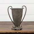 The 11.5" ESPERANZA VASE by Ragon House, featuring a rustic, weathered metal finish and two curved handles, sits on a wooden surface in front of a white shiplap wall.