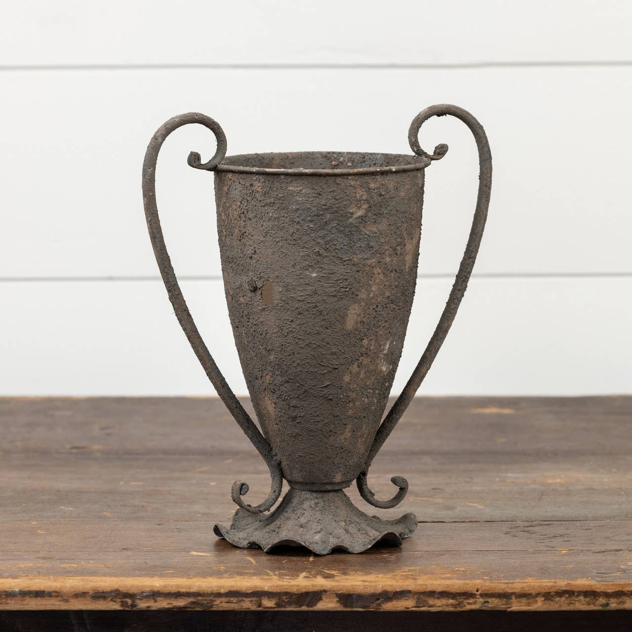 The 11.5" ESPERANZA VASE by Ragon House, featuring a rustic, weathered metal finish and two curved handles, sits on a wooden surface in front of a white shiplap wall.