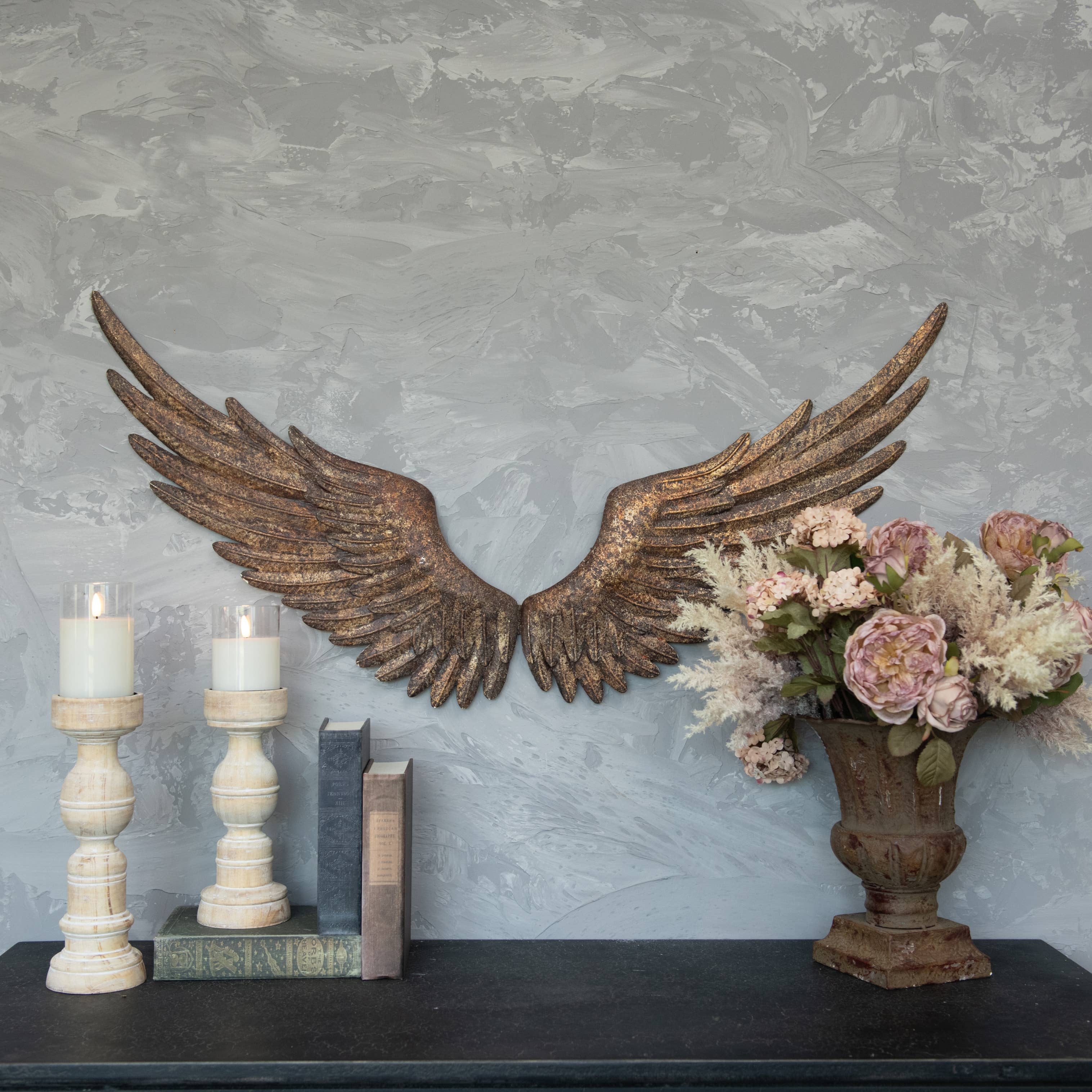 A decorative wall features Ragon House's ANTIQUE REPRODUCTION GOLD WALL WINGS mounted with keyhole hangers, accented by two white candle holders, three stacked books, and a vase of pink and white flowers on a dark tabletop.