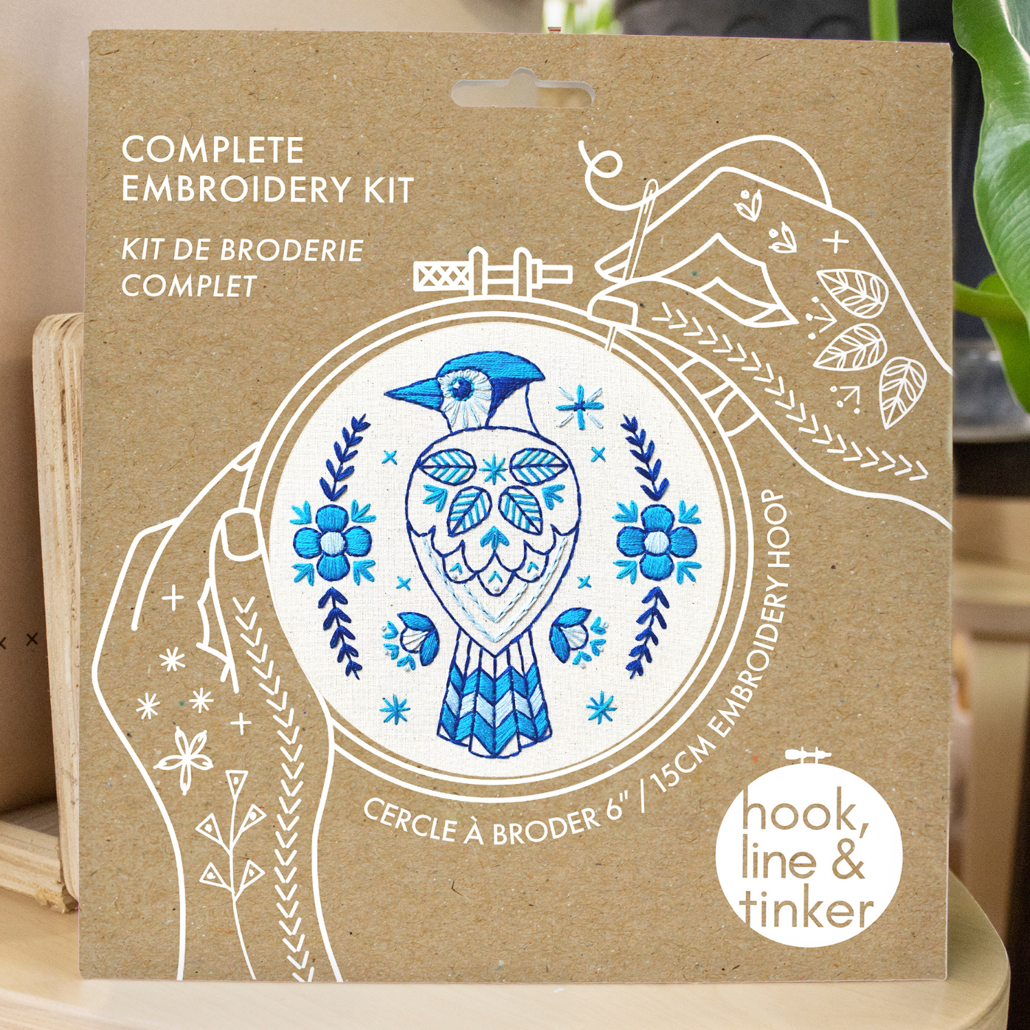 The Blue Jay Complete Embroidery Kit by Hook, Line & Tinker Embroidery Kits comes in a cardboard package featuring a blue jay and floral hoop design, plus white line drawings of hands stitching on the box.