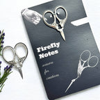 A black Firefly Notes notebook labeled "Petite Thread Snips" rests on a white surface, next to ornate vintage-style sewing scissors and purple flowers. The notebook cover features an illustration of small scissors.