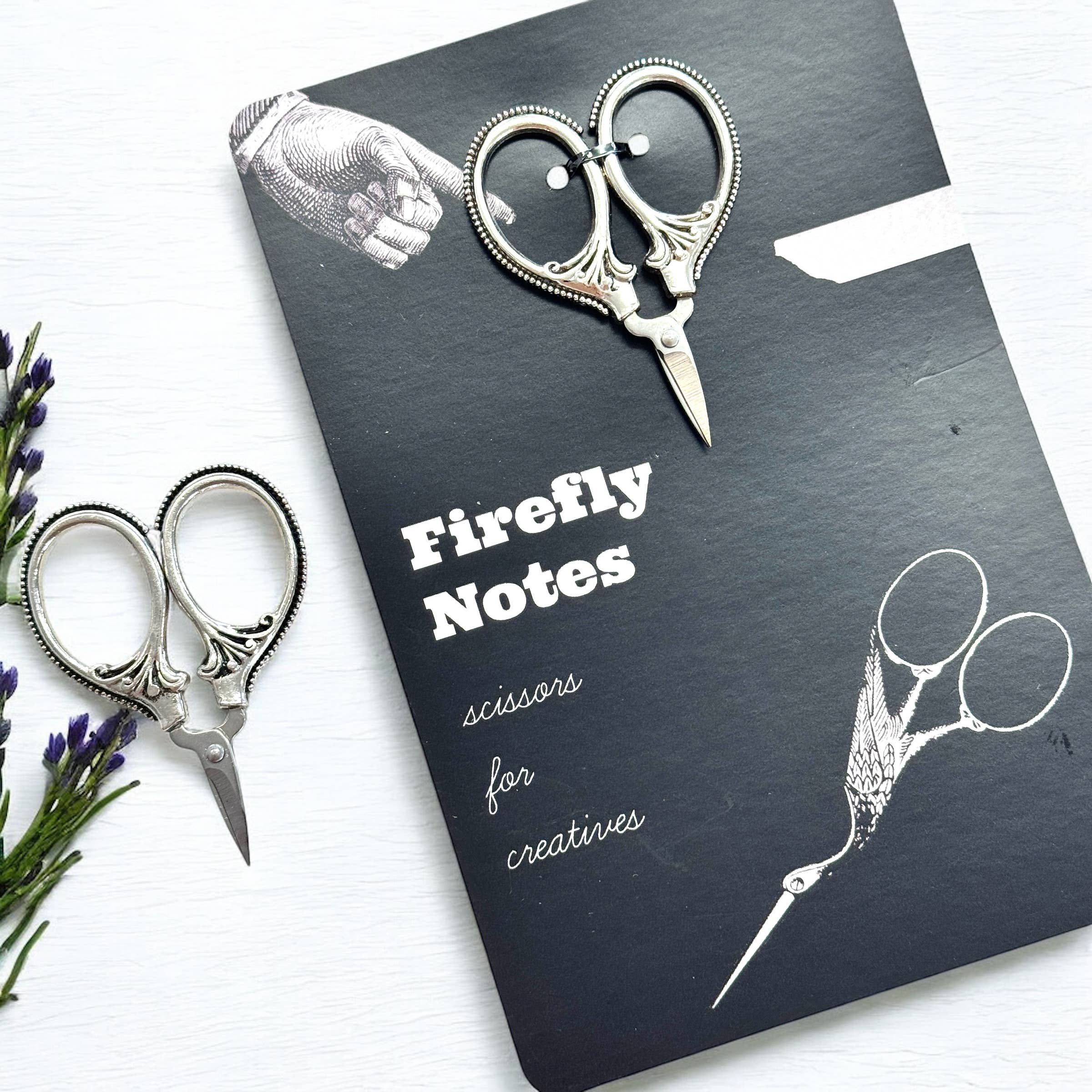 A black Firefly Notes notebook labeled "Petite Thread Snips" rests on a white surface, next to ornate vintage-style sewing scissors and purple flowers. The notebook cover features an illustration of small scissors.