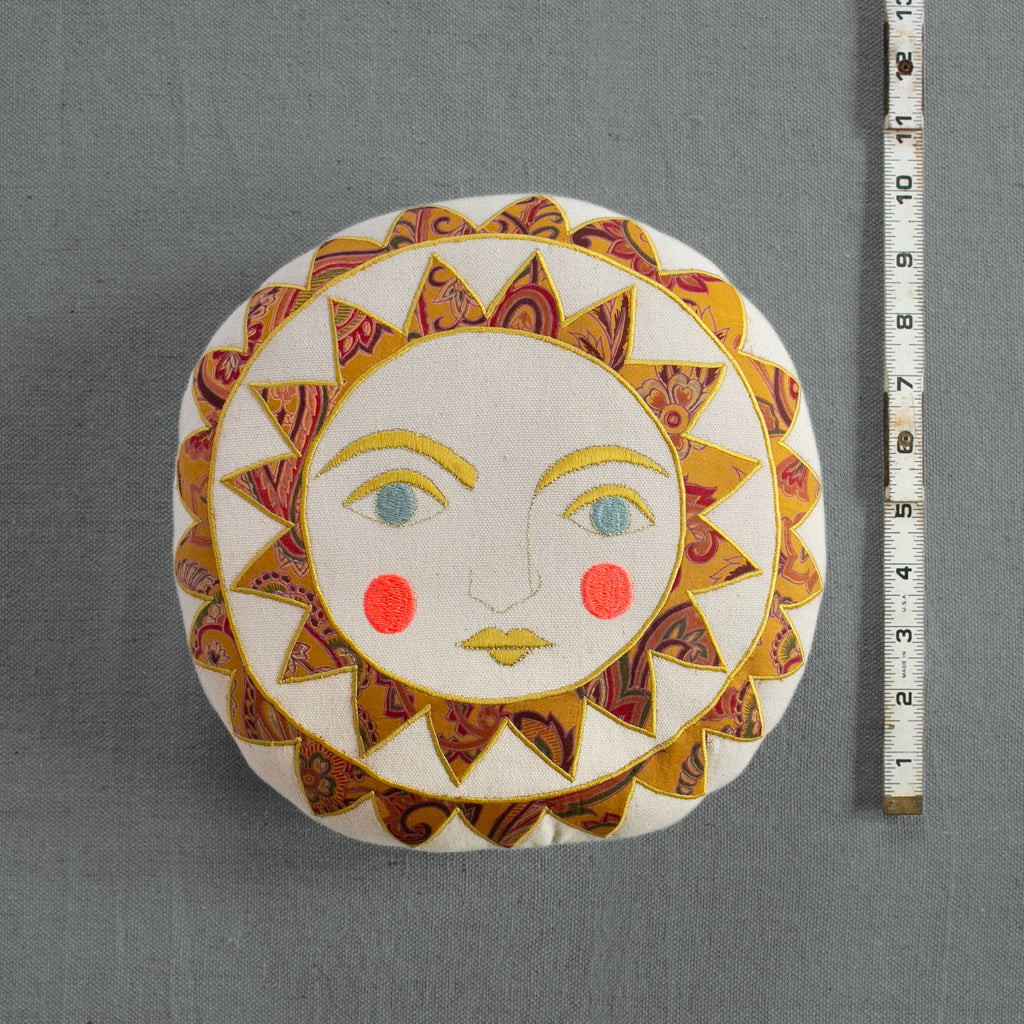 The Skippy Cotton Bright Neon Sun Pillow is a round, cotton-filled, handmade cushion with a whimsical embroidered sun face—yellow, red, and patterned rays, blue eyes, and rosy cheeks. A measuring tape sits vertically beside it on gray fabric.