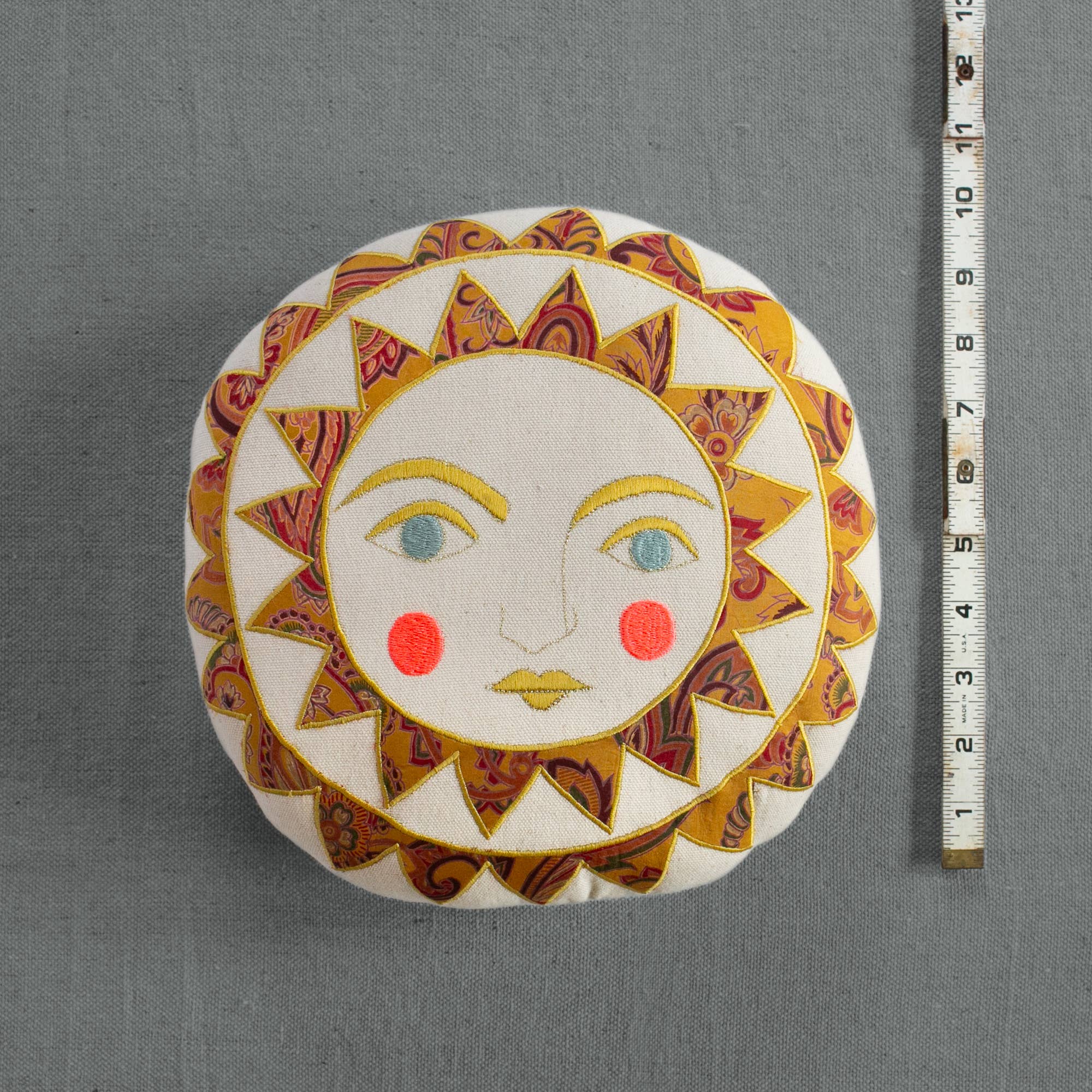 The Skippy Cotton Bright Neon Sun Pillow is a round, cotton-filled, handmade cushion with a whimsical embroidered sun face—yellow, red, and patterned rays, blue eyes, and rosy cheeks. A measuring tape sits vertically beside it on gray fabric.