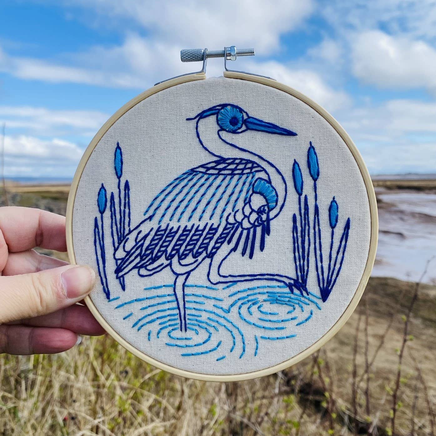 A hand holds the Blue Heron Complete Embroidery Kit by Hook, Line & Tinker Embroidery Kits, featuring a blue heron among cattails stitched in blue thread—perfect for beginner crafters inspired by nature.