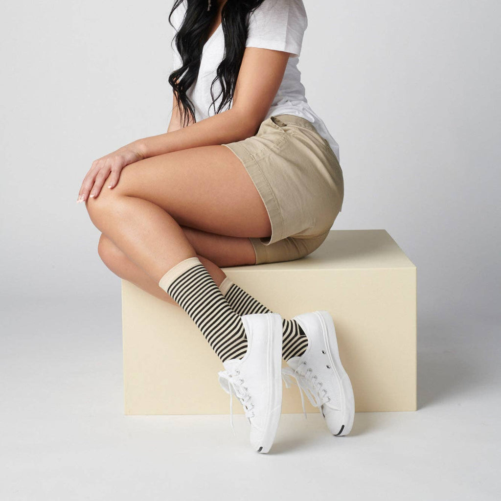 A person sits on a light block wearing a white t-shirt, khaki shorts, Hooray Sock Co. Cole Black and White Striped Crew Socks, and white sneakers. The background is plain and light; the person’s face is not shown.