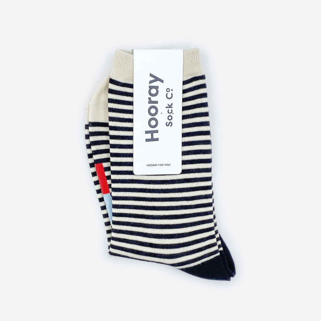 The Cole Black and White Striped Crew Socks by Hooray Sock Co. feature crew length, cream-colored toes and heels, and come partially wrapped in a white label with the brand’s name in large gray letters for unisex comfort and style.