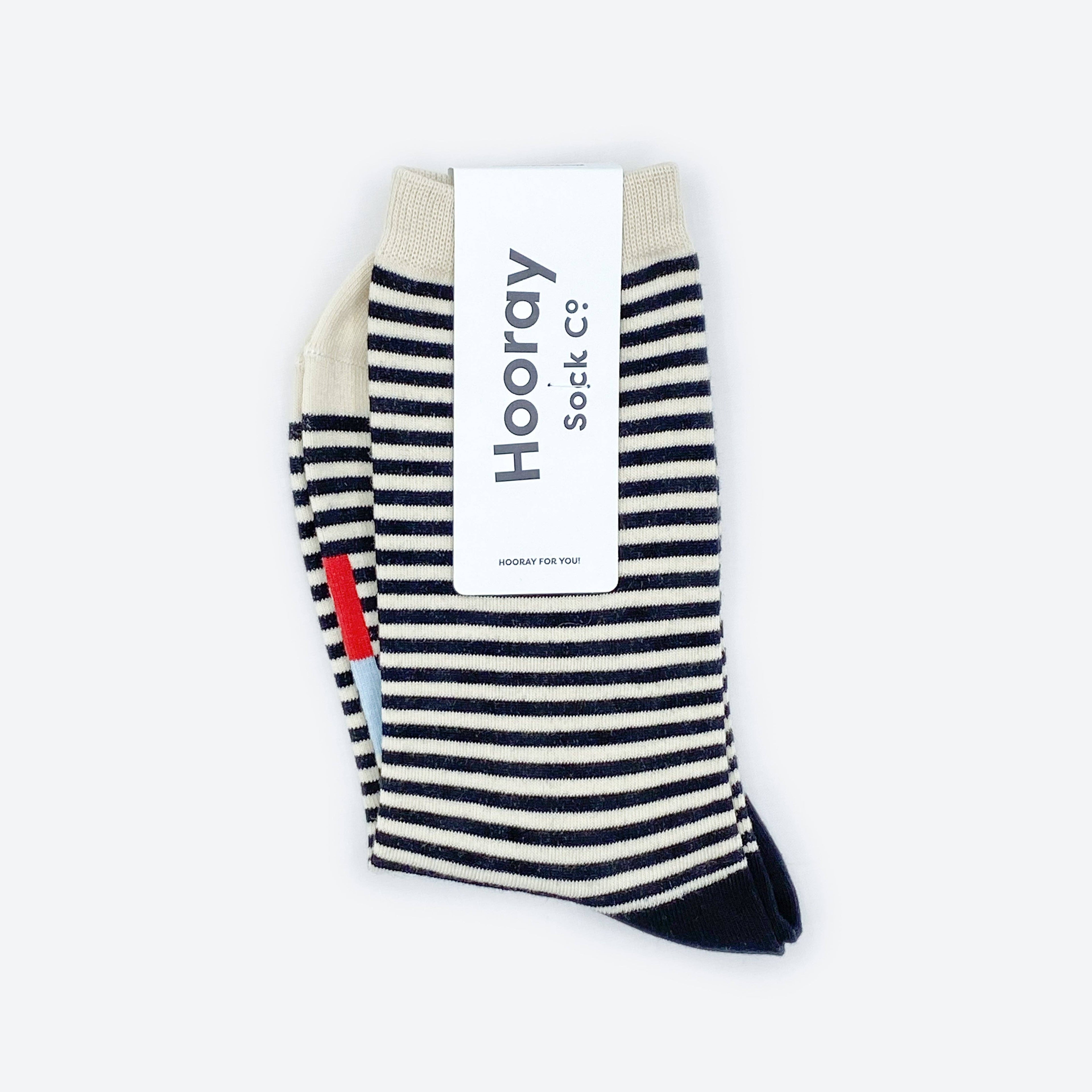 The Cole Black and White Striped Crew Socks by Hooray Sock Co. feature crew length, cream-colored toes and heels, and come partially wrapped in a white label with the brand’s name in large gray letters for unisex comfort and style.