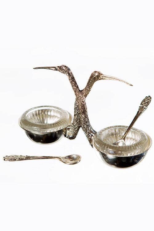 The Stork Salt and Pepper Cellar by Vagabond Vintage features two silver bowls with glass inserts and matching spoons, joined by back-to-back stork figures with long necks and beaks for a decorative touch.