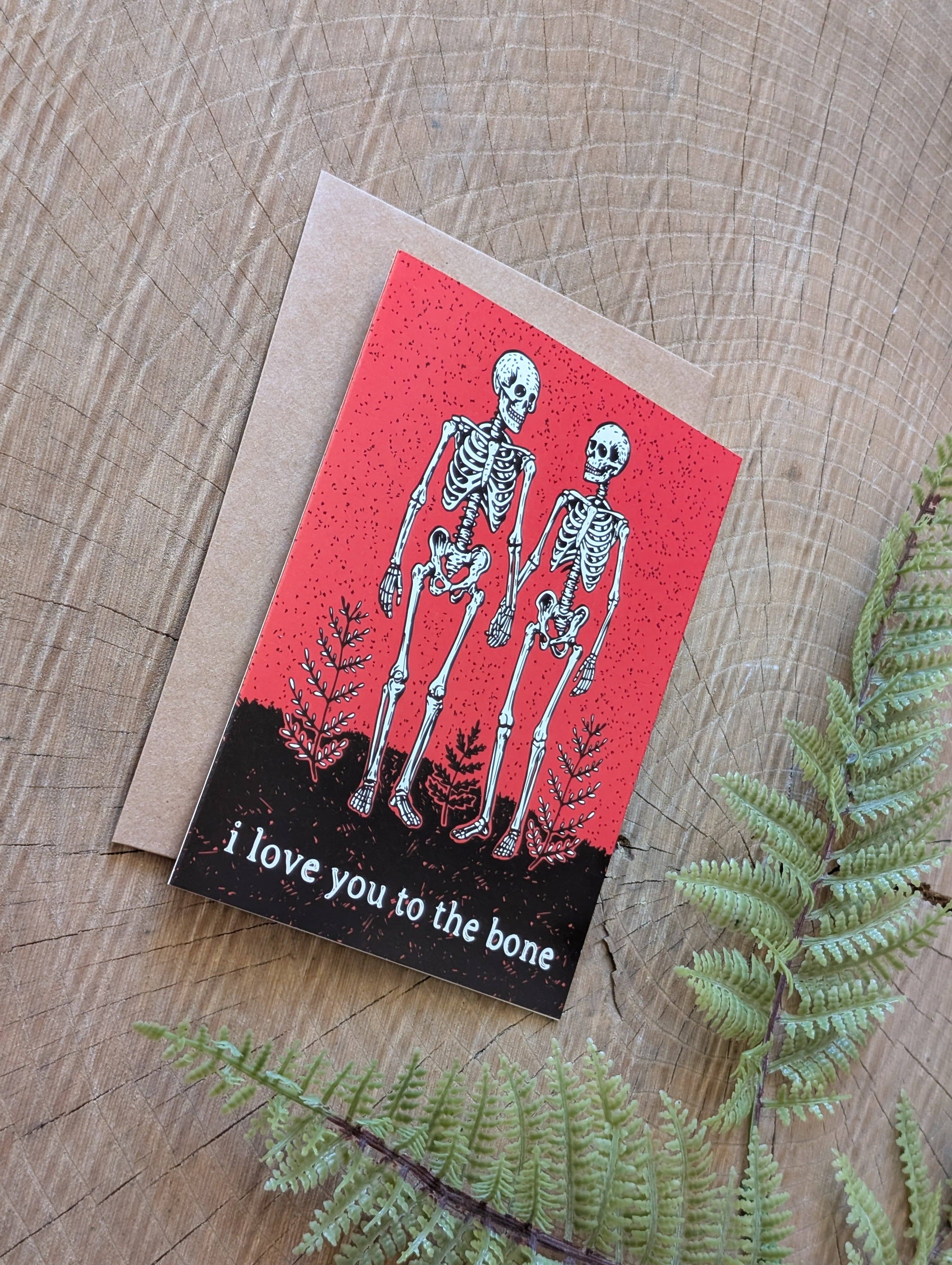 The 'i love you to the bone' greeting card by ilikesara | art + goods features two skeletons holding hands on brown paper with red and fern accents—a romantic touch, perfect for loved ones.