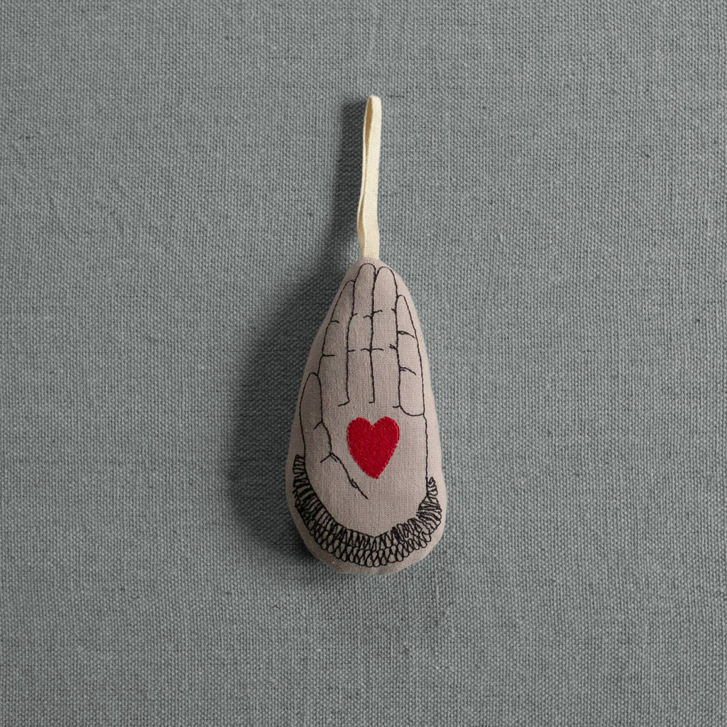 The Skippy Cotton Heart in Hand - Cotton & Lavender filled Ornament features a teardrop shape with a black hand and red heart design on organic cotton, hanging by a loop against a gray textured background.