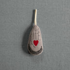 The Skippy Cotton Heart in Hand - Cotton & Lavender filled Ornament features a teardrop shape with a black hand and red heart design on organic cotton, hanging by a loop against a gray textured background.