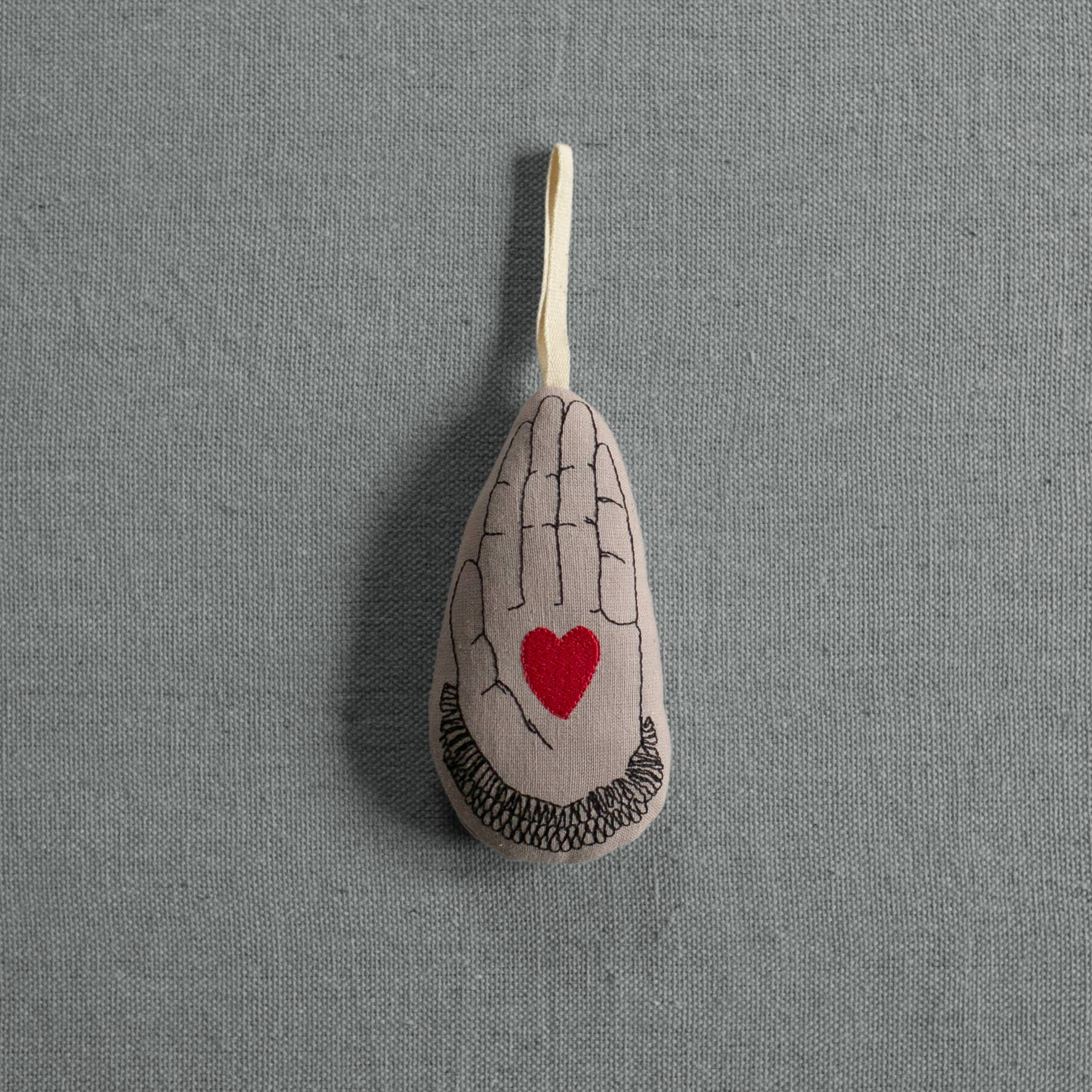 The Skippy Cotton Heart in Hand - Cotton & Lavender filled Ornament features a teardrop shape with a black hand and red heart design on organic cotton, hanging by a loop against a gray textured background.