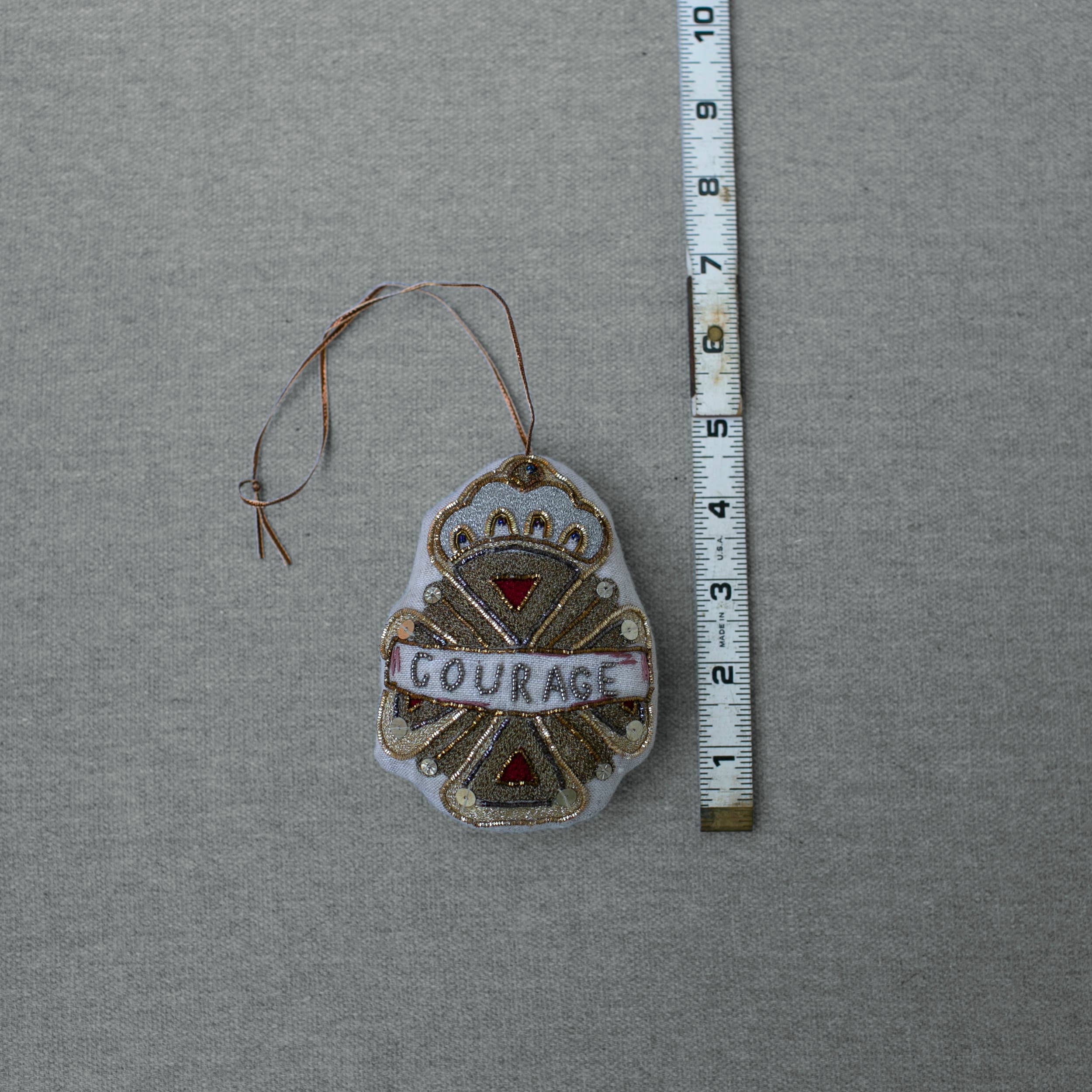 The Skippy Cotton Courage Crest Token, an embellished Oz cotton-filled ornament with gold and red accents and "COURAGE" at its center, measures about 3 inches long and is shown on a grey background beside a measuring tape.