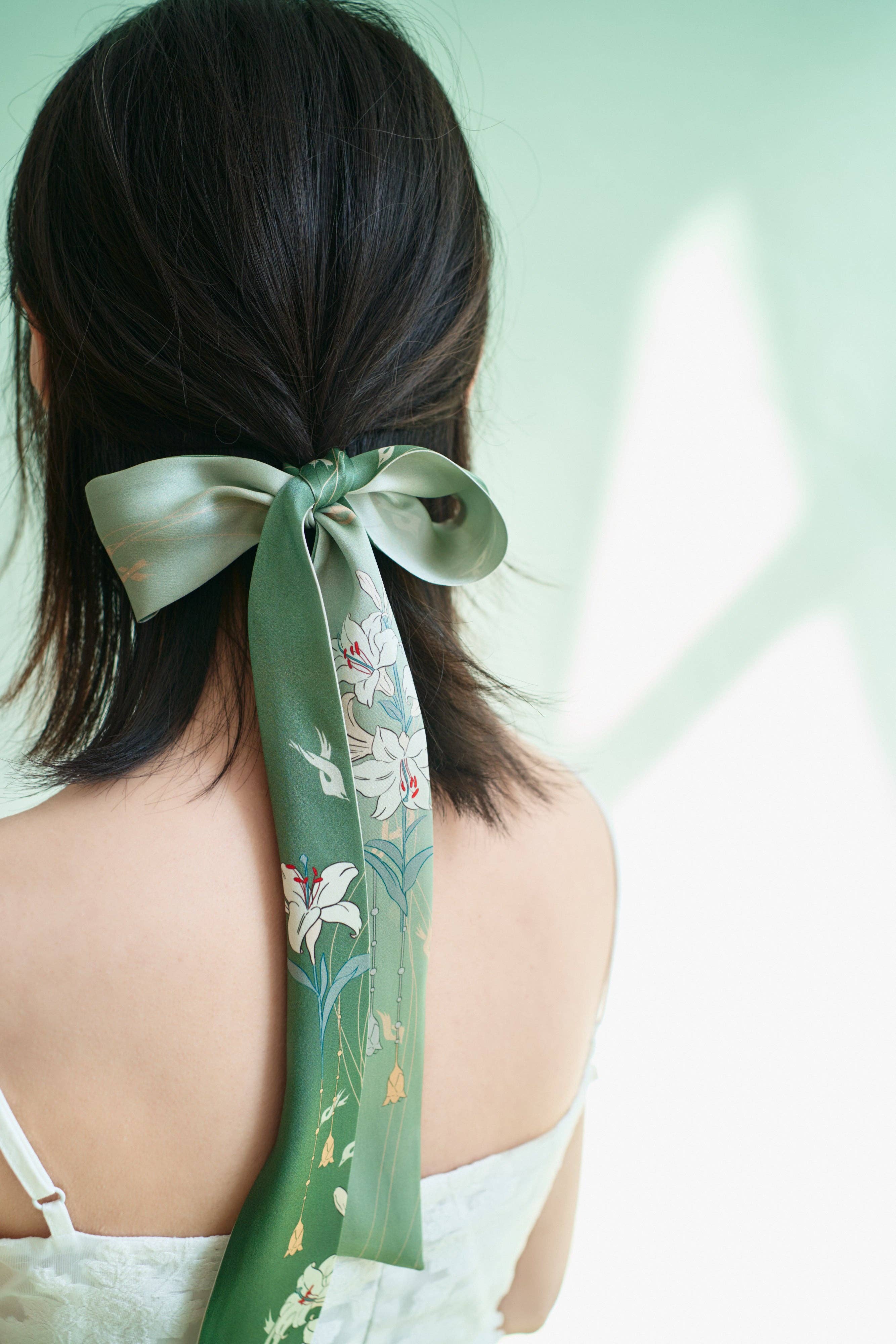 A woman with shoulder-length dark hair wears the Lost Pattern NYC Lily Silk Twilly Scarf in Green, tied in a bow at the back of her head. She faces away from the camera, dressed in a white sleeveless top, against a soft green background.