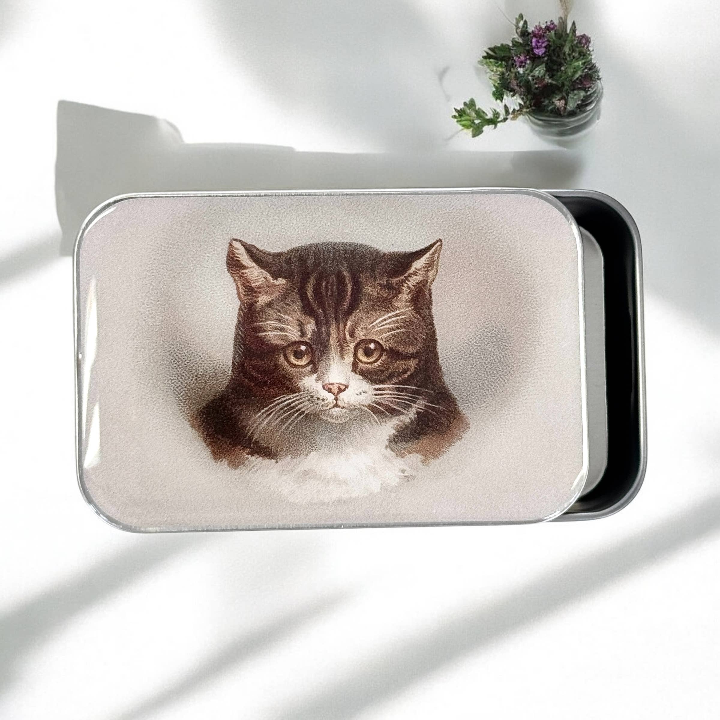A small, handcrafted Firefly Notes Cat Notions Tin with a resin-coated finish features Moody the Cat’s face in a realistic illustration on the lid, shown on a white surface beside flowers, its durable gloss catching soft shadows.