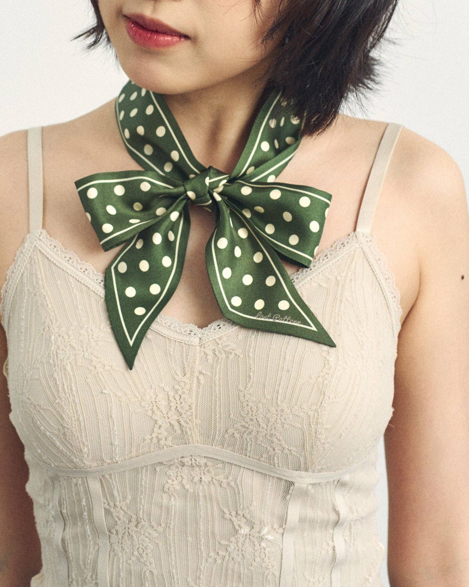 A woman with short dark hair wears a beige lace camisole and the Lost Pattern NYC Silk Skinny Scarf in Dark Green, tied in a bow around her neck. Her face is partially visible.