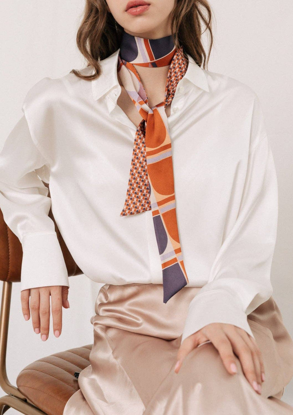 A woman wears the Lost Pattern NYC Silk Skinny Scarf in Clay tied around her neck with a white satin blouse and beige skirt, adding a chic touch to her wavy hair as a stylish accessory while she sits on a brown chair.