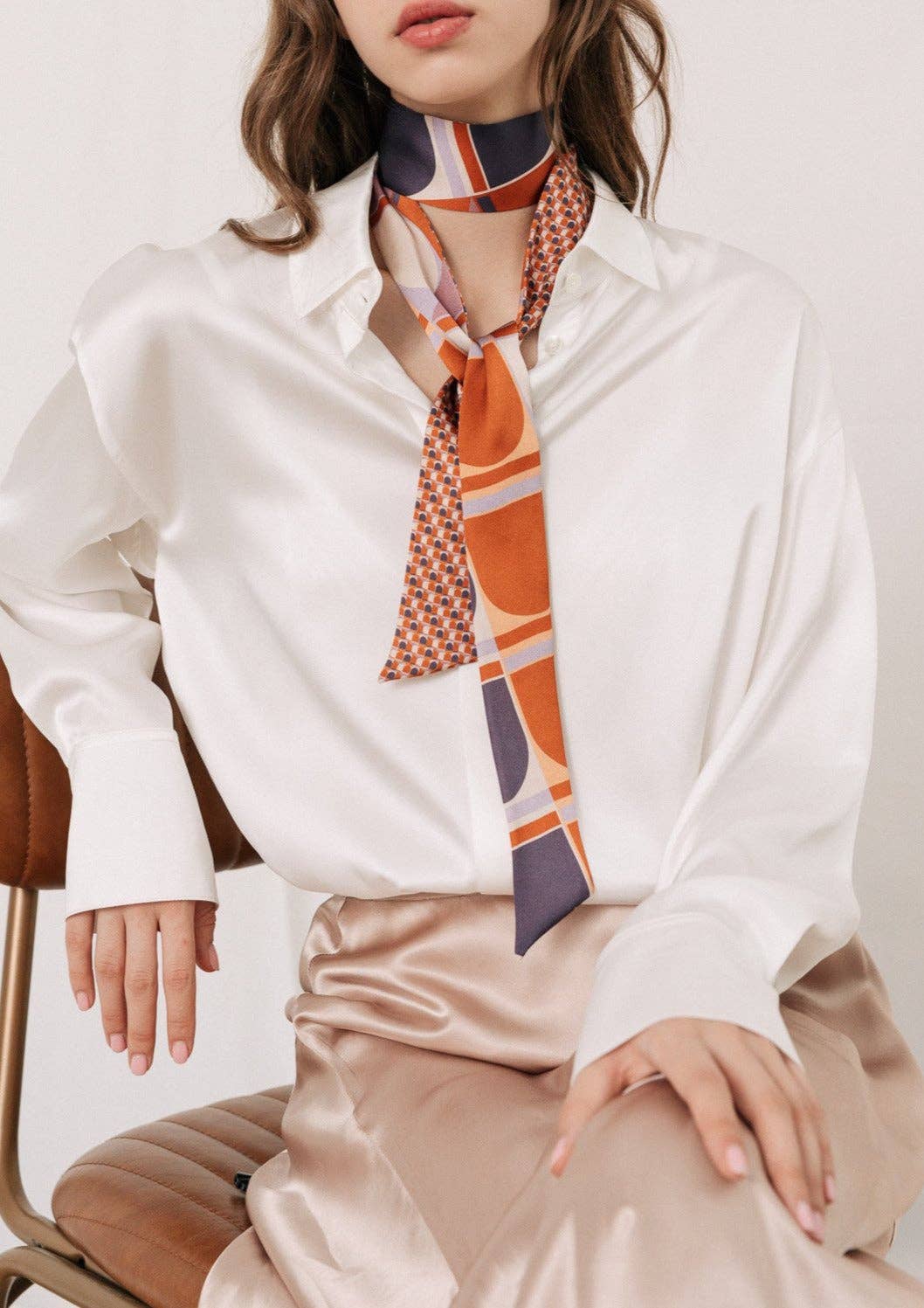 A woman wears the Lost Pattern NYC Silk Skinny Scarf in Clay tied around her neck with a white satin blouse and beige skirt, adding a chic touch to her wavy hair as a stylish accessory while she sits on a brown chair.