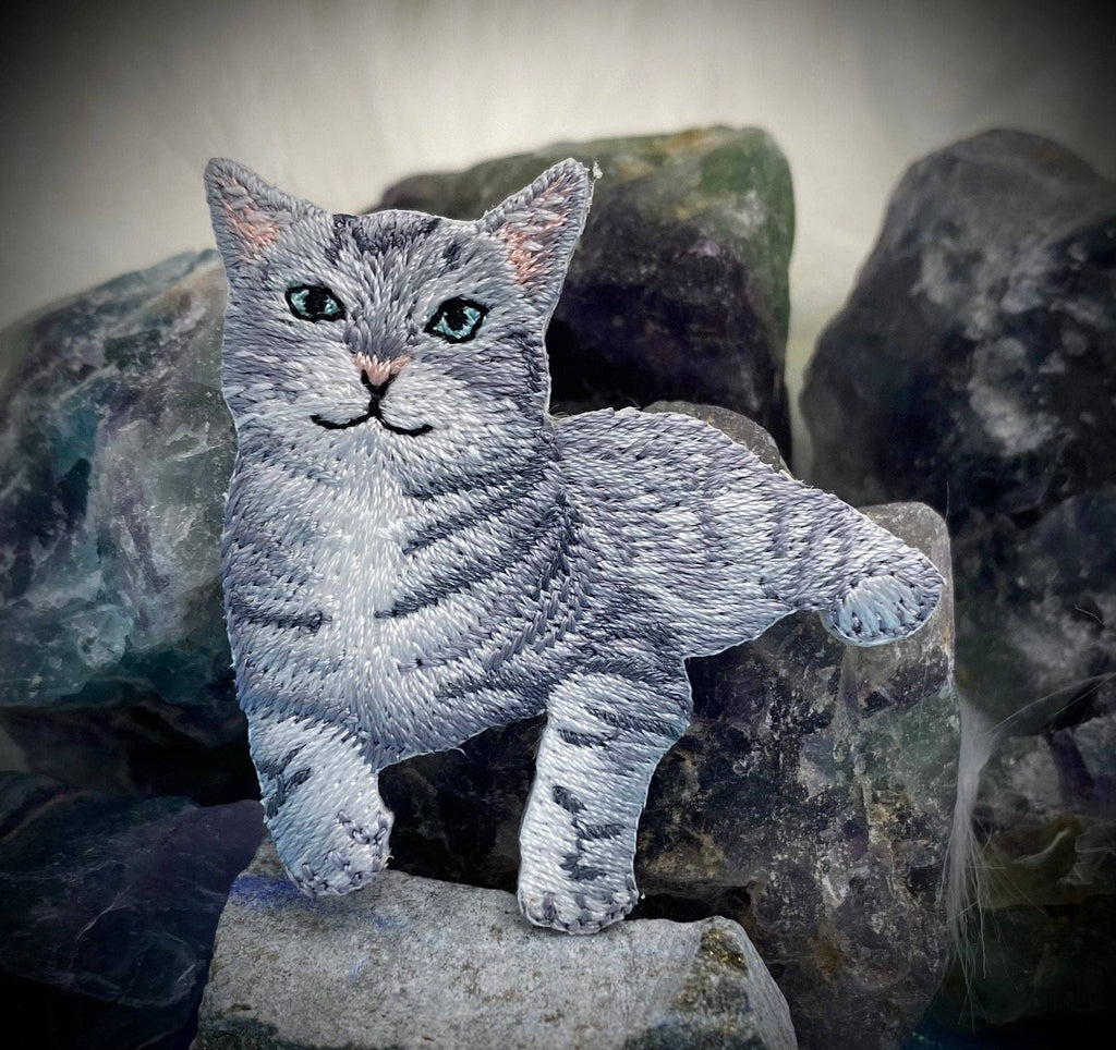 The Wholesale Applique Gray Tabby Cat Iron on Patch, featuring a green-eyed kitten design, is pictured atop rough multicolored stones with a blurred white background—perfect for adding charm to any fabric project.