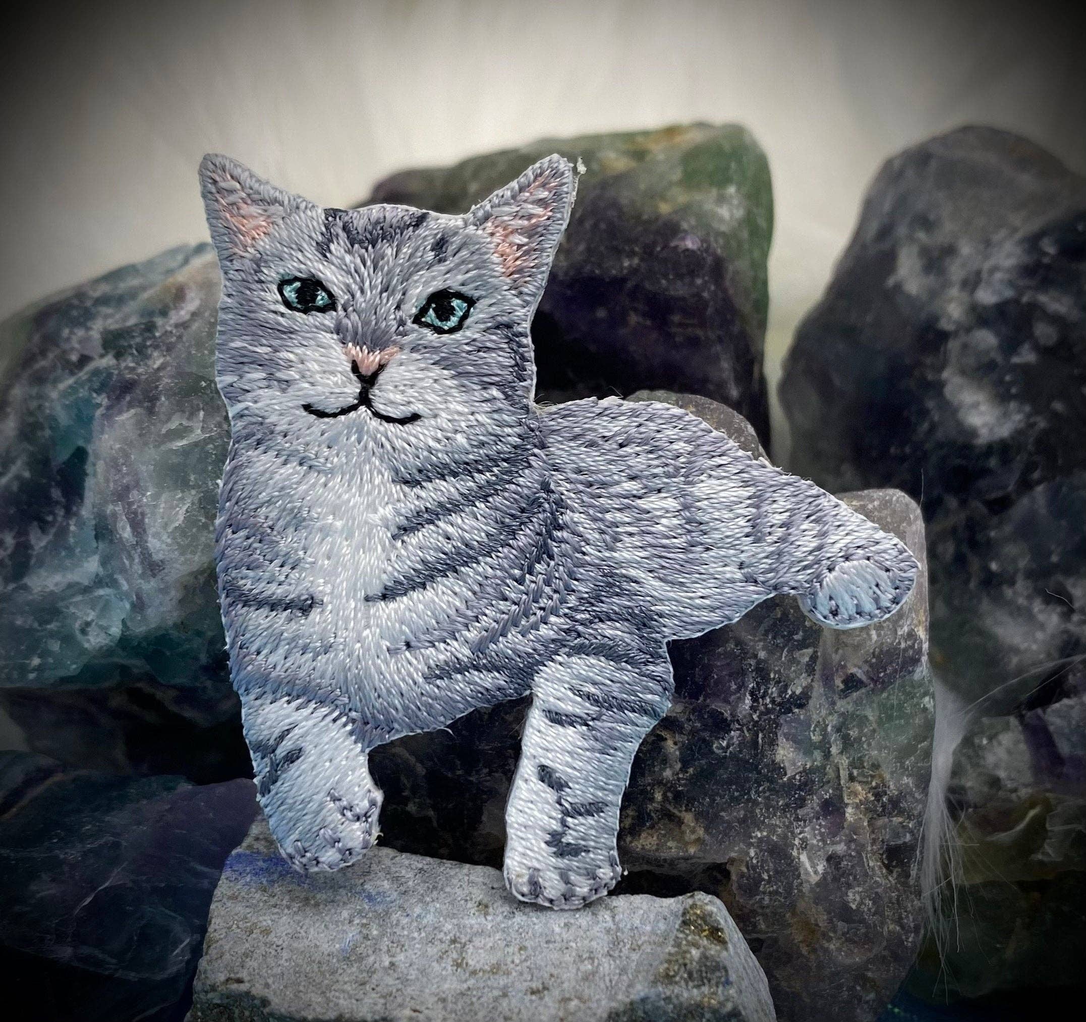 The Wholesale Applique Gray Tabby Cat Iron on Patch, featuring a green-eyed kitten design, is pictured atop rough multicolored stones with a blurred white background—perfect for adding charm to any fabric project.
