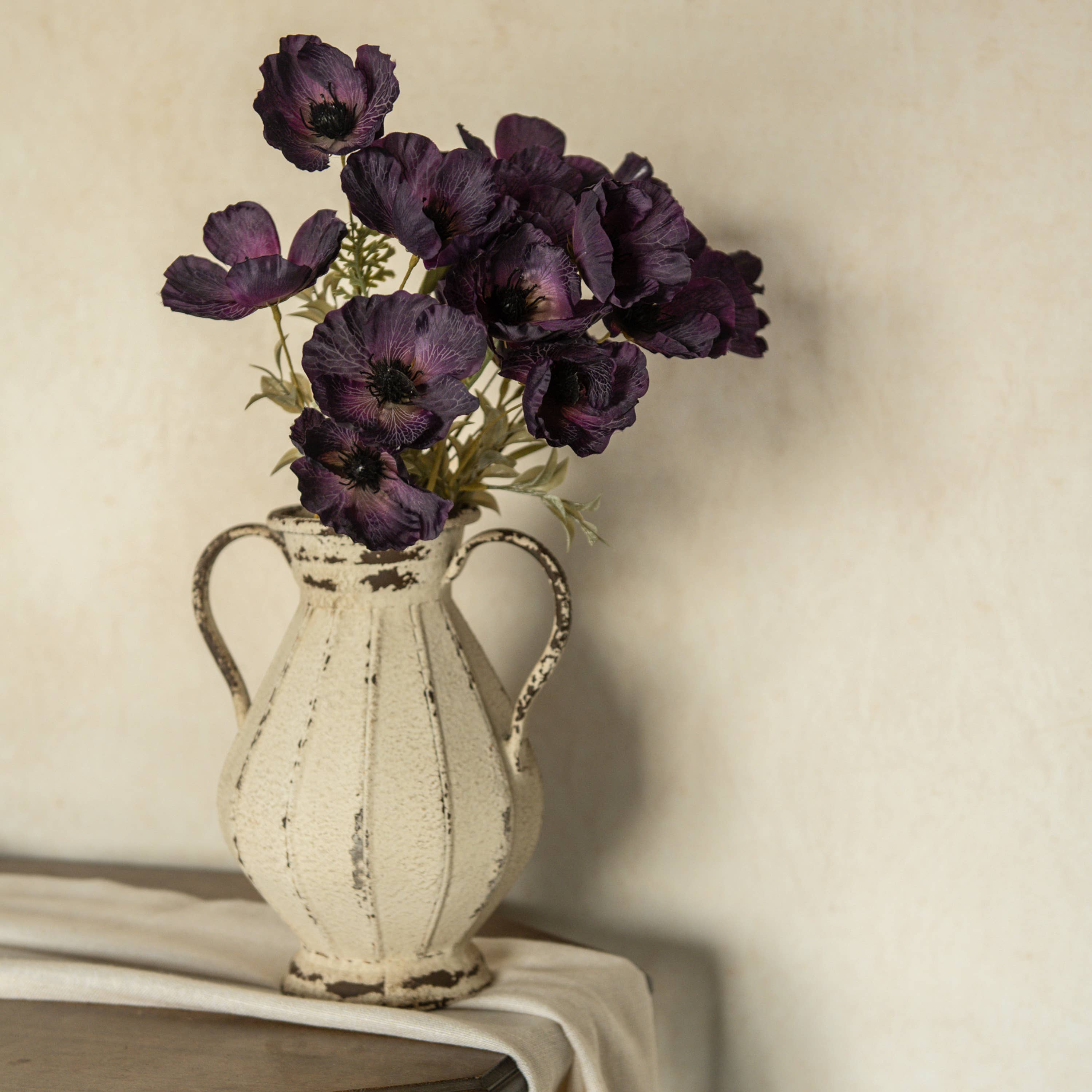 A vintage cream vase holds the 22" Artificial Dark Purple Poppy Bush by Ragon House, displayed on draped fabric with a neutral textured background.