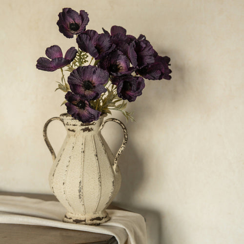 A vintage cream vase holds the 22" Artificial Dark Purple Poppy Bush by Ragon House, displayed on draped fabric with a neutral textured background.