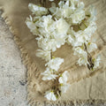 The FAUX WHITE CHERRY BLOSSOM SPRAY by Ragon House features delicate white blossoms atop a beige fringed fabric, placed on textured stone for a soft, rustic look.