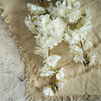 The FAUX WHITE CHERRY BLOSSOM SPRAY by Ragon House features delicate white blossoms atop a beige fringed fabric, placed on textured stone for a soft, rustic look.