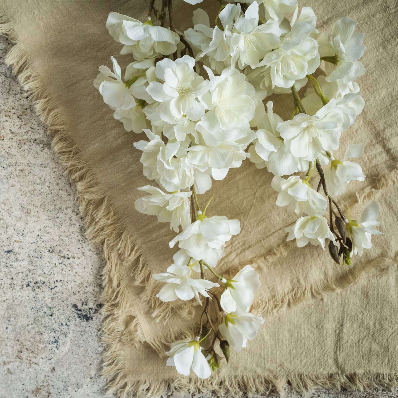 The FAUX WHITE CHERRY BLOSSOM SPRAY by Ragon House features delicate white blossoms atop a beige fringed fabric, placed on textured stone for a soft, rustic look.