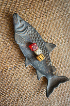 The Vagabond Vintage Cast Iron Fish Dish with a rust finish holds three dice—red, white, and yellow—on a woven, textured surface.