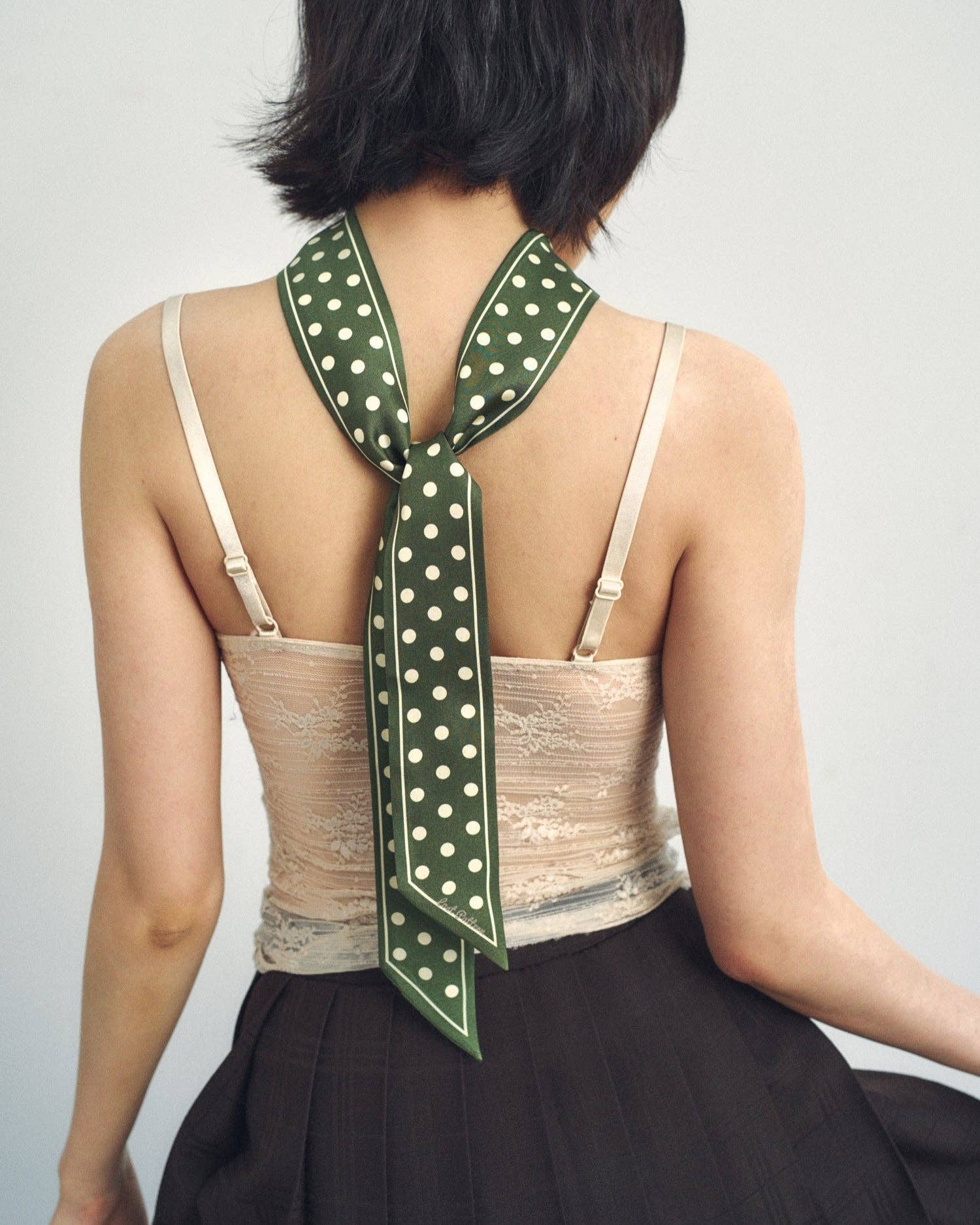 A woman with short dark hair, seen from behind, wears a lace camisole, a dark pleated skirt, and the Lost Pattern NYC Silk Skinny Scarf in Dark Green tied around her neck with long ends draping down her back.