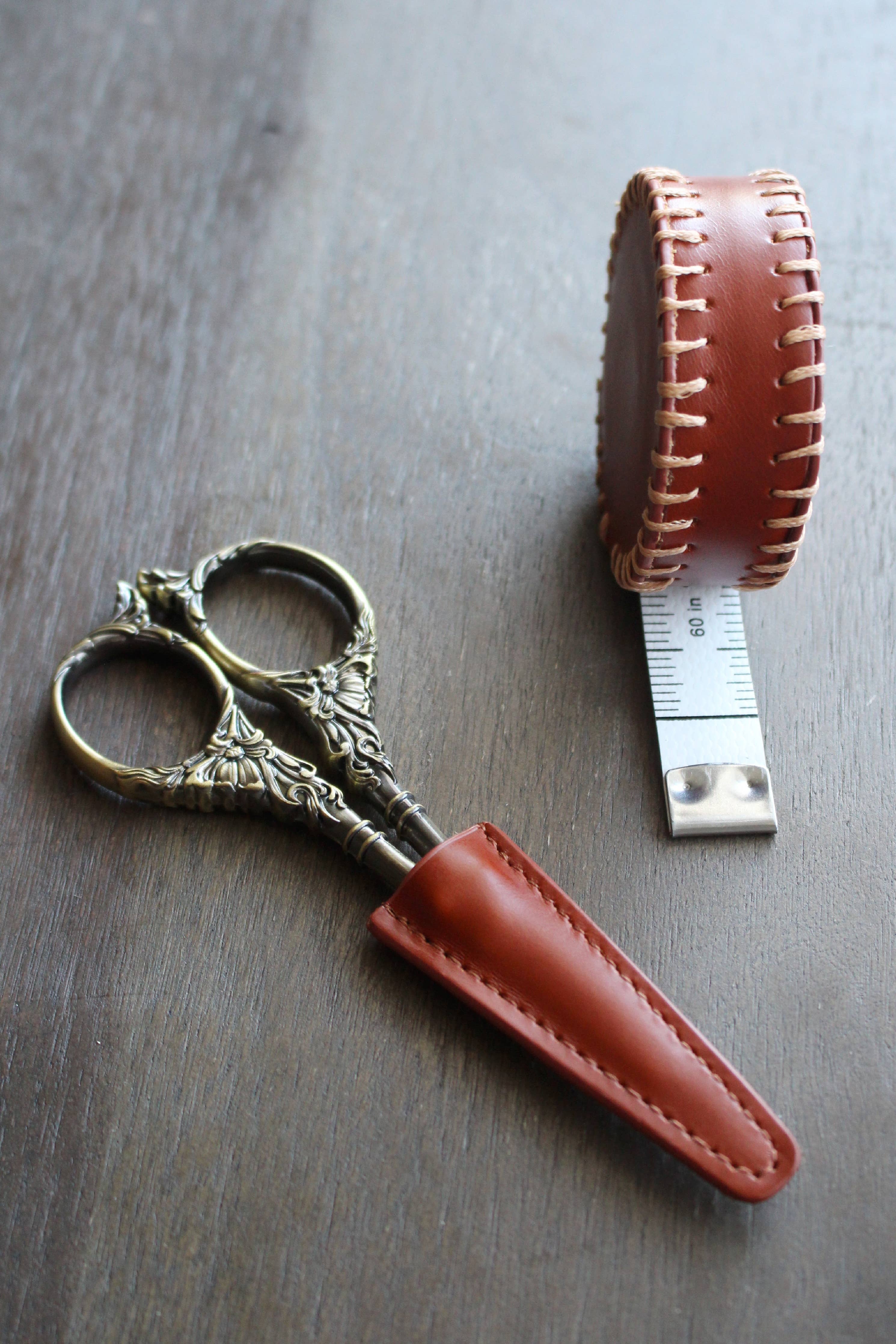 A pair of ornate embroidery scissors in an NNK Press Leather Scissor Sheath rests on a wooden surface beside a round, brown leather tape measure with the measuring tape partly extended.