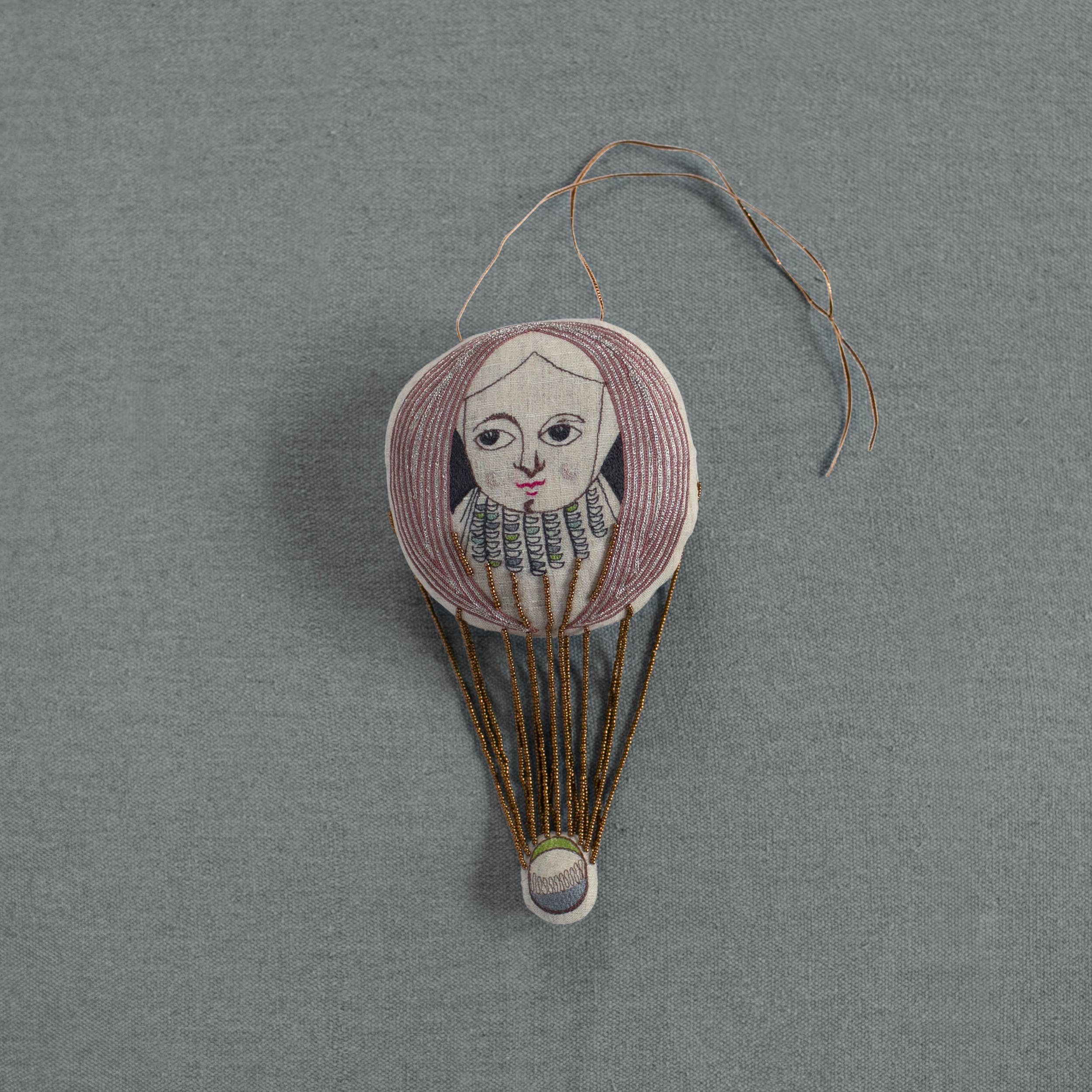 The Skippy Cotton "Wonderful Wizard / Balloon" wallhanging ornament features a hand-stitched face with pink hair, ruffled collar, and gold threads on a gray background, creating whimsical embroidered art inspired by Oz.