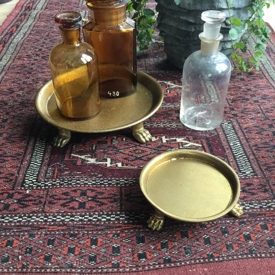 Two amber and one clear glass bottles sit on a patterned rug with a Vagabond Vintage Small Claw Foot Round Dish with Gold Leaf: Large holding the bottles, while another dish stands empty. Greenery accents the background.