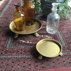 Two amber and one clear glass bottles sit on a patterned rug with a Vagabond Vintage Small Claw Foot Round Dish with Gold Leaf: Large holding the bottles, while another dish stands empty. Greenery accents the background.