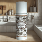 A bottle of Roots And Leaves Botanical Magnesium Deodorant sits on a kitchen counter with a tiled backsplash, sink, and utensils in the background. The label displays botanical illustrations.