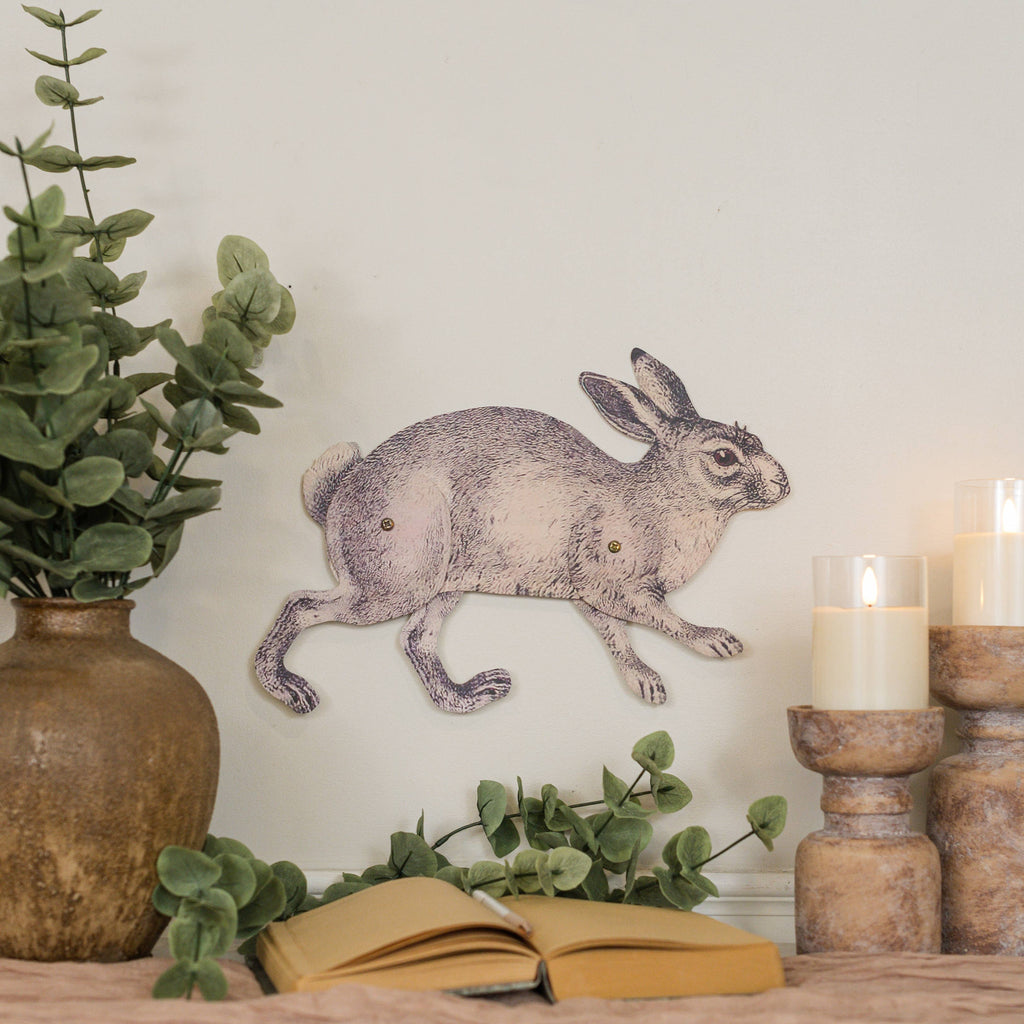 The Ragon House POSEABLE METAL BUNNY, featuring movable legs, hangs on the wall above a table with 14-inch hangers. Below are green foliage, a brown vase, lit candles on wooden stands, and an open book.