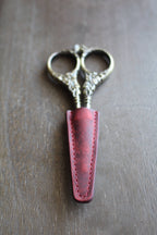 A pair of vintage embroidery scissors, partly covered by the NNK Press Leather Scissor Sheath, rest on a wooden surface.