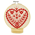 The Nordic Heart Complete Embroidery Kit by Hook, Line & Tinker features a red heart with floral and leaf accents on cream fabric atop a wooden base—ideal for beginner embroiderers.
