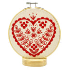 The Nordic Heart Complete Embroidery Kit by Hook, Line & Tinker features a red heart with floral and leaf accents on cream fabric atop a wooden base—ideal for beginner embroiderers.