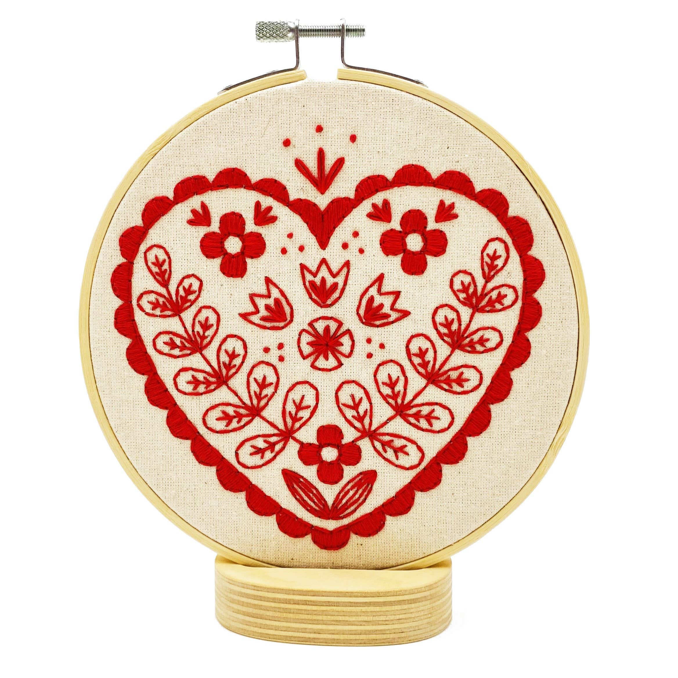 The Nordic Heart Complete Embroidery Kit by Hook, Line & Tinker features a red heart with floral and leaf accents on cream fabric atop a wooden base—ideal for beginner embroiderers.