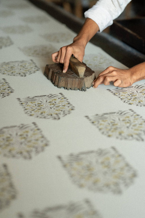 A person demonstrates traditional block printing techniques by pressing a wooden block onto fabric, creating intricate patterns on the Indaba Trading Botanique Block Print Tablecloth Pink. Hands and part of the patterned textile are in focus.
