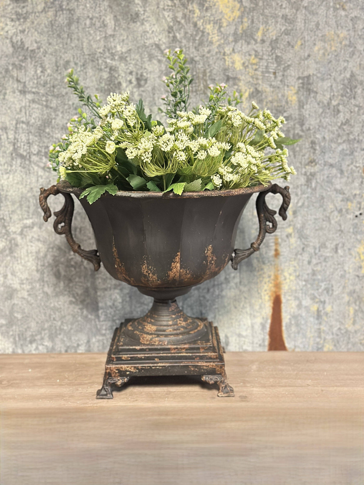 The Distressed Black Iron Urn by Vagabond Vintage, featuring ornate handles, displays white flowers and green foliage on a wooden surface against a weathered wall.