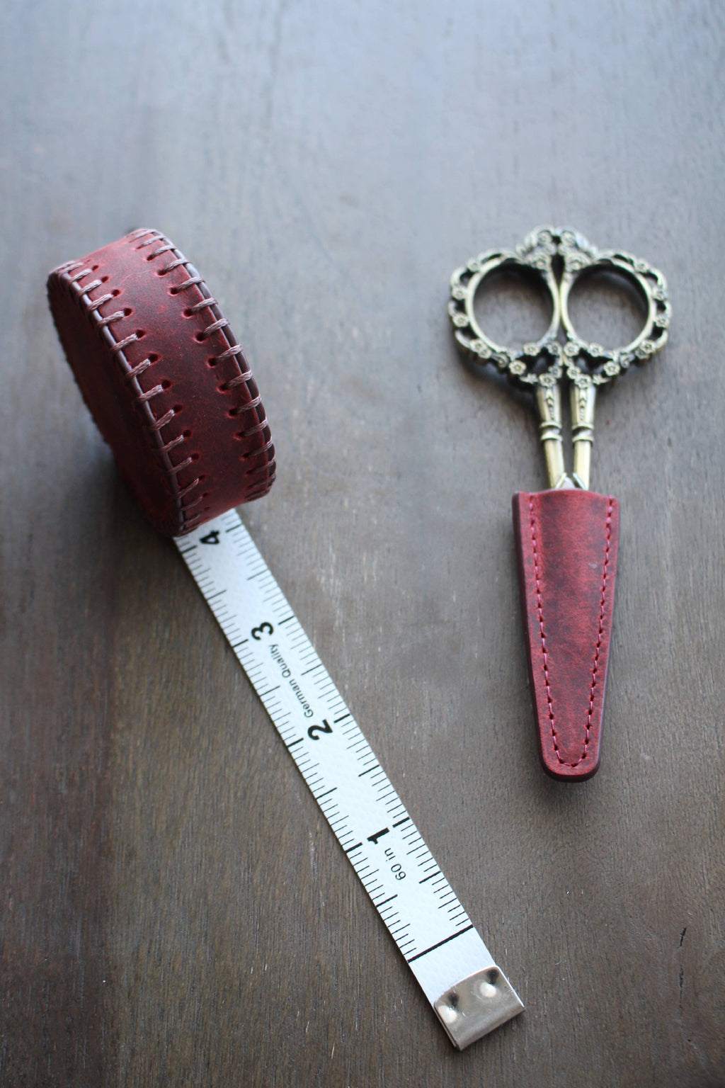 A metal tape measure with a red leather case and ornate embroidery scissors in an NNK Press Leather Scissor Sheath rest on a wooden surface.